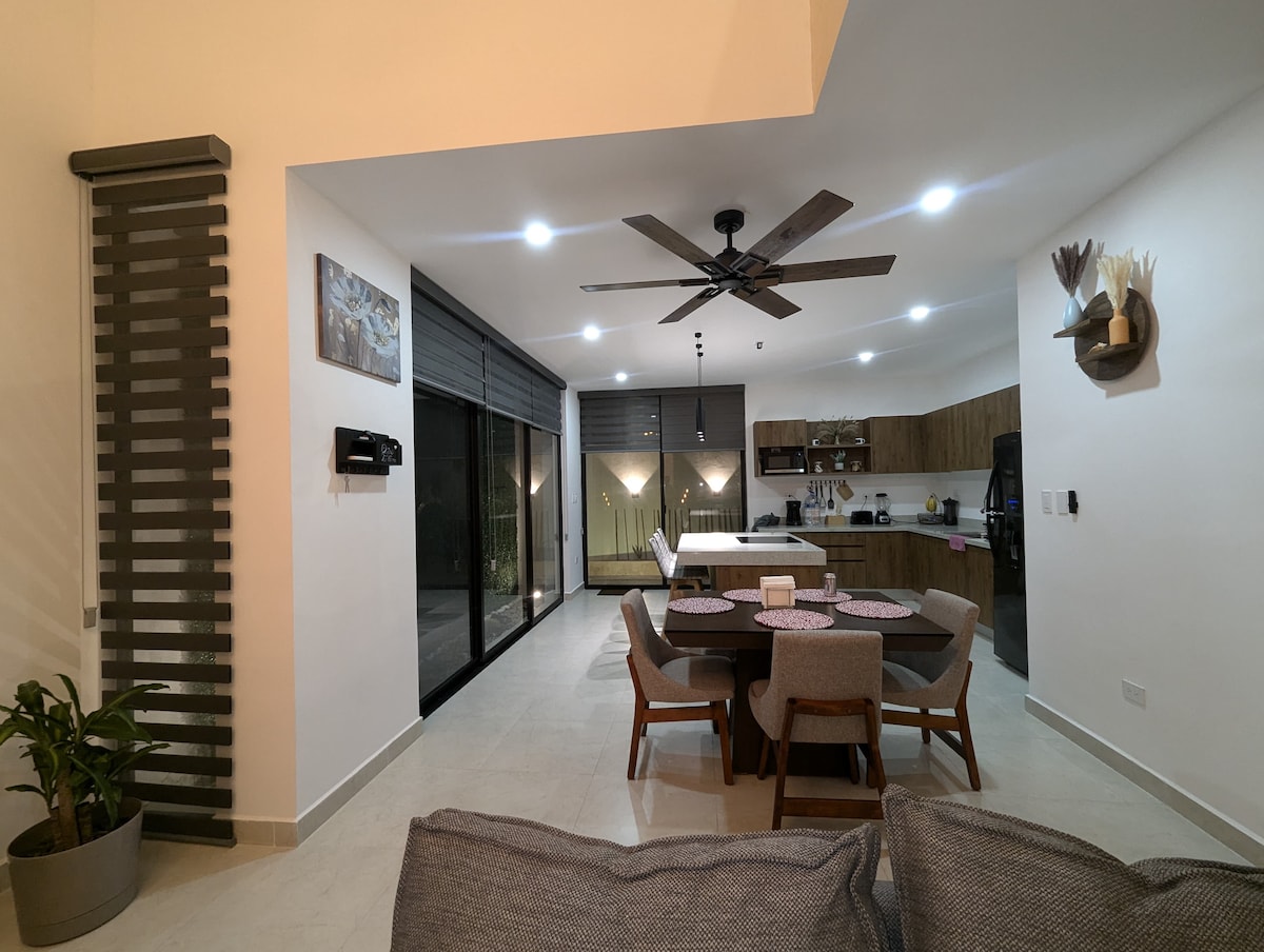 The open-plan living area is illuminated by recessed lighting, with a ceiling fan overhead. A comfortable sofa is visible alongside a round dining table set for four. The kitchen, featuring wooden cabinetry and modern appliances, is accessed through expansive glass doors that lead to an outdoor space.