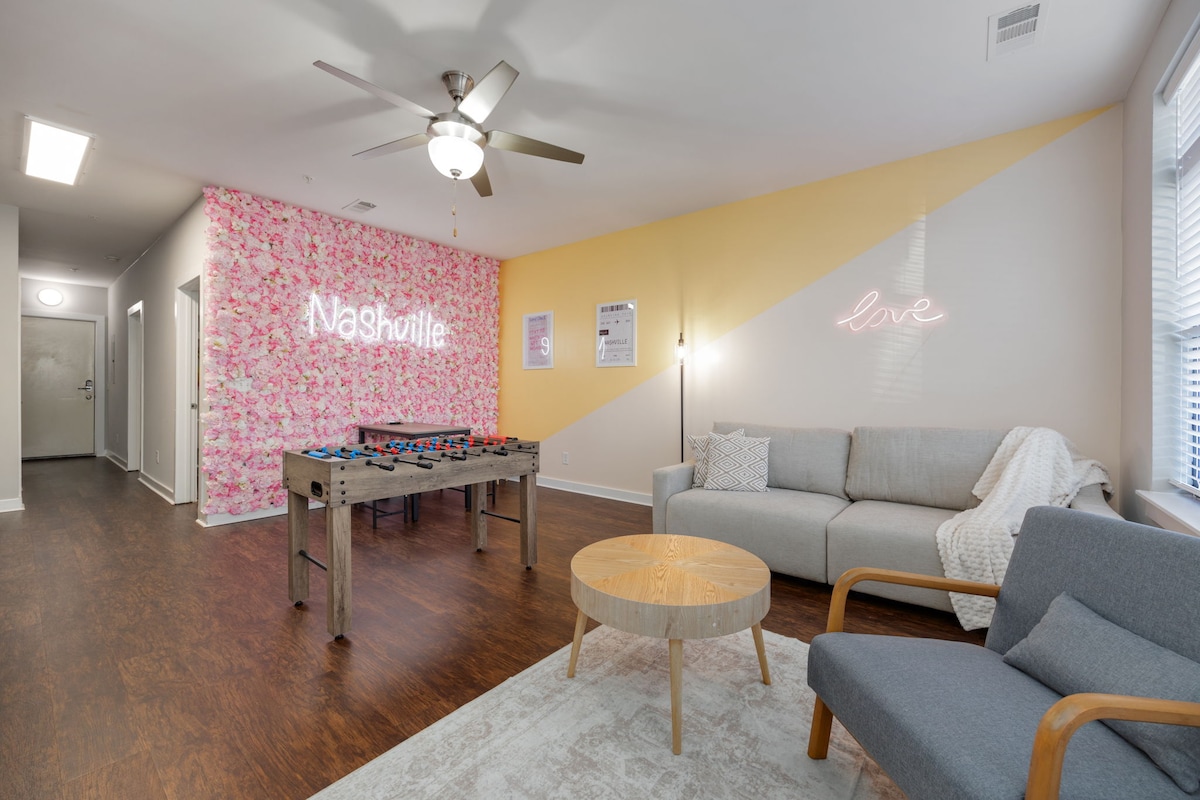 A welcoming living area features a foosball table and a comfortable couch, with a round coffee table at the center. Vibrant floral wallpaper on one wall complements a soft pastel accent, while a neon sign displays 'Nashville' and another states 'love.'