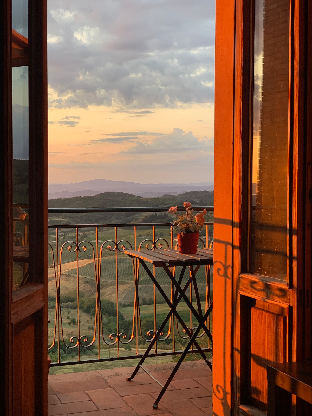 A balcony door opens to reveal stunning views of rolling hills at sunset. A small table holds a potted flower, enhancing the tranquil setting. The warm glow of the evening light bathes the wooden door frame, creating a welcoming ambiance.