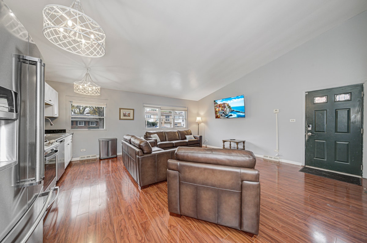 The bright living area features modern lighting fixtures and hardwood flooring. A spacious seating arrangement includes a stylish brown leather sofa and armchairs, facing a smart TV mounted on the wall. A front door adds access to the outdoor space, enhancing the open feel.