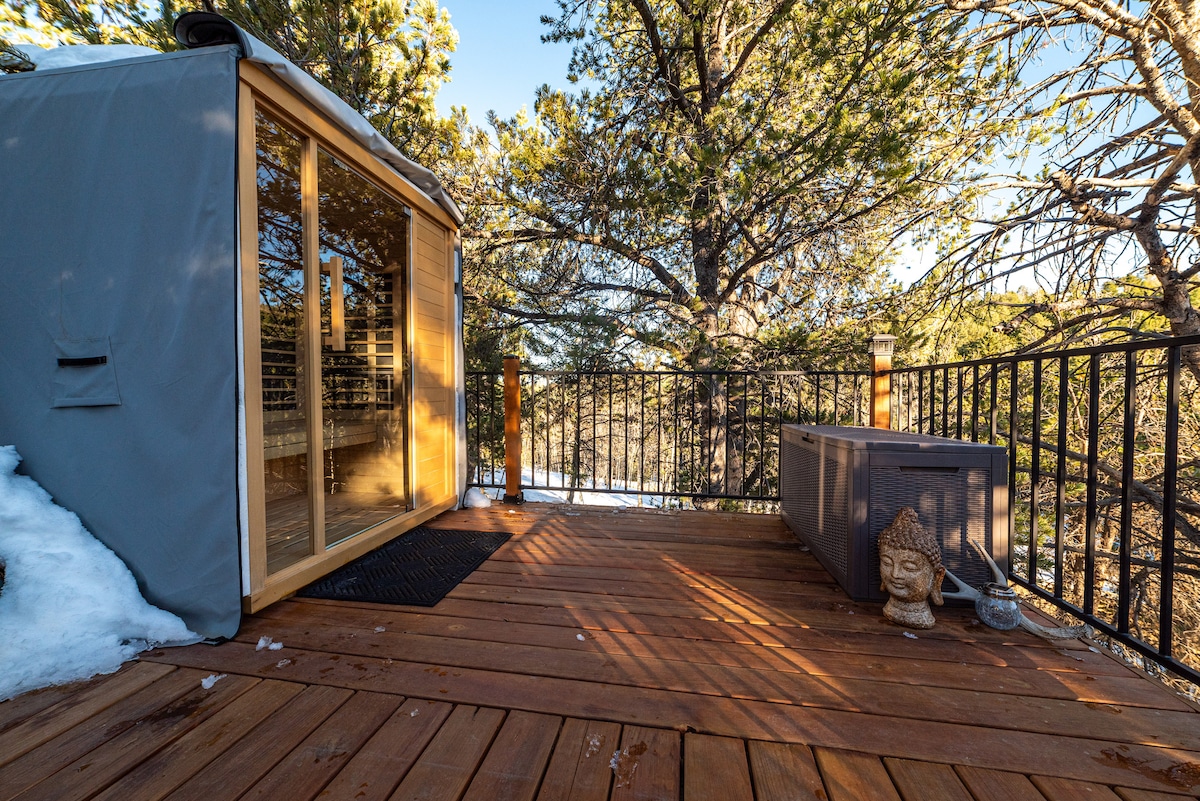 A spacious wooden deck offers a relaxing outdoor space surrounded by trees. An infrared sauna is positioned on the left, while a propane fire pit is located near the right side, creating a cozy atmosphere. Snow coverage can be seen beside the deck.