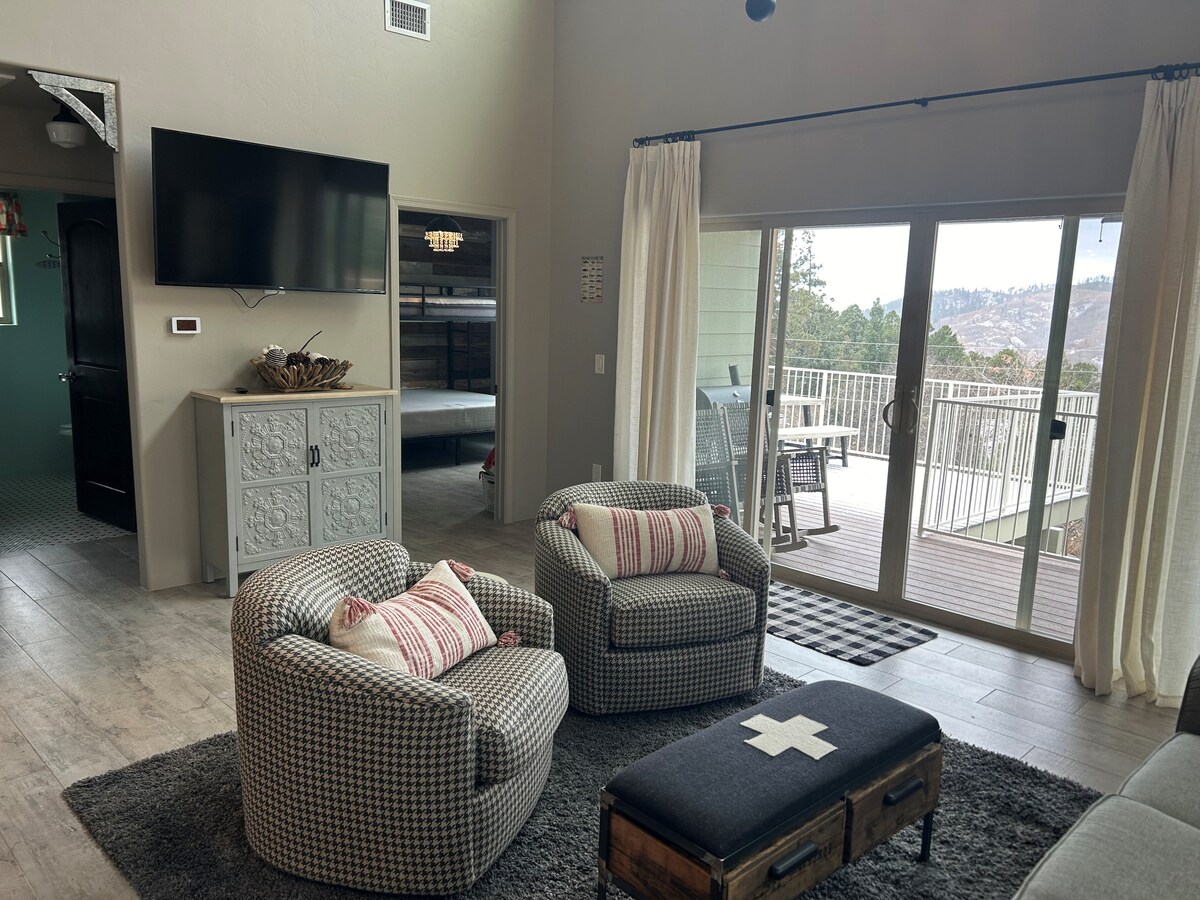 A welcoming living area is highlighted by two patterned armchairs and a coffee table with a cross design. A large television is mounted on the wall, and natural light streams through sliding glass doors that open to a deck with mountain views.