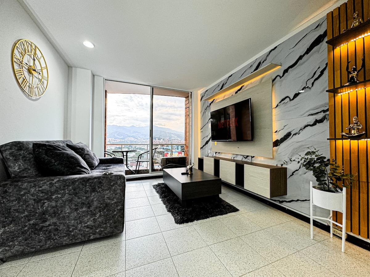 A modern living area is presented with a dark sofa and a marble accent wall. A flat-screen TV is mounted on the wall, and a glass sliding door offers access to a balcony with city views. Natural light brightens the room, enhancing the spacious atmosphere.