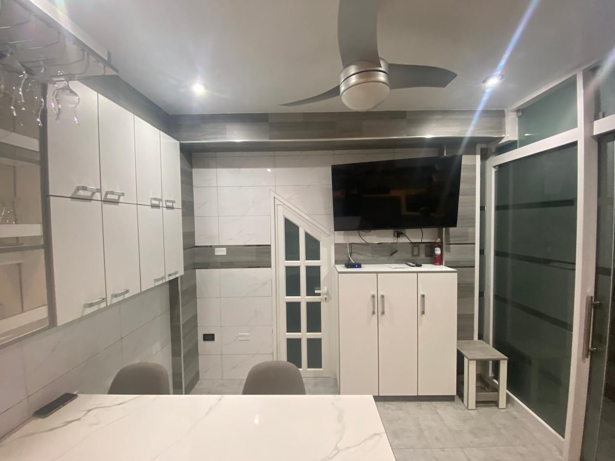 A modern kitchen area features sleek cabinets and a large dining table. A mounted television is seen against a decorative wall, with a ceiling fan above providing air circulation. The space is complemented by glass doors that lead to a separate area.