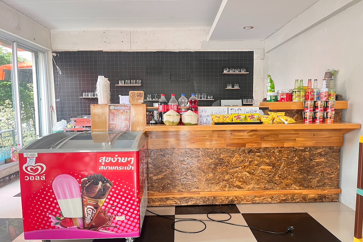 A counter area features a wooden bar top with various beverages displayed in glass bottles. A freezer holds ice cream products, while shelves are stocked with snacks and drinks. The space is bright and inviting, with large windows allowing natural light to enter.