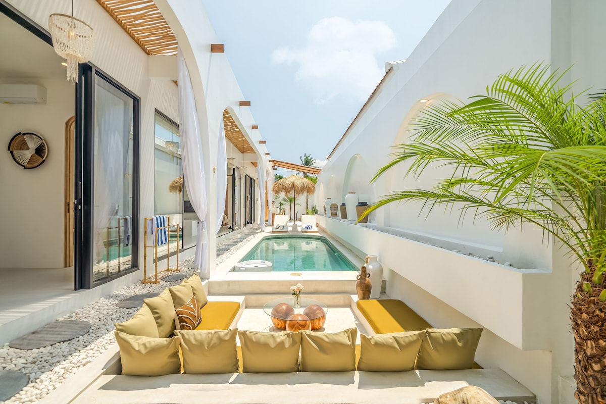An outdoor seating area is framed by lush greenery, featuring comfortable cushions arranged around a glass coffee table. The serene pool reflects the clear sky, while palm trees add to the tropical ambiance. Arched structures and light-colored walls create a spacious, inviting setting.