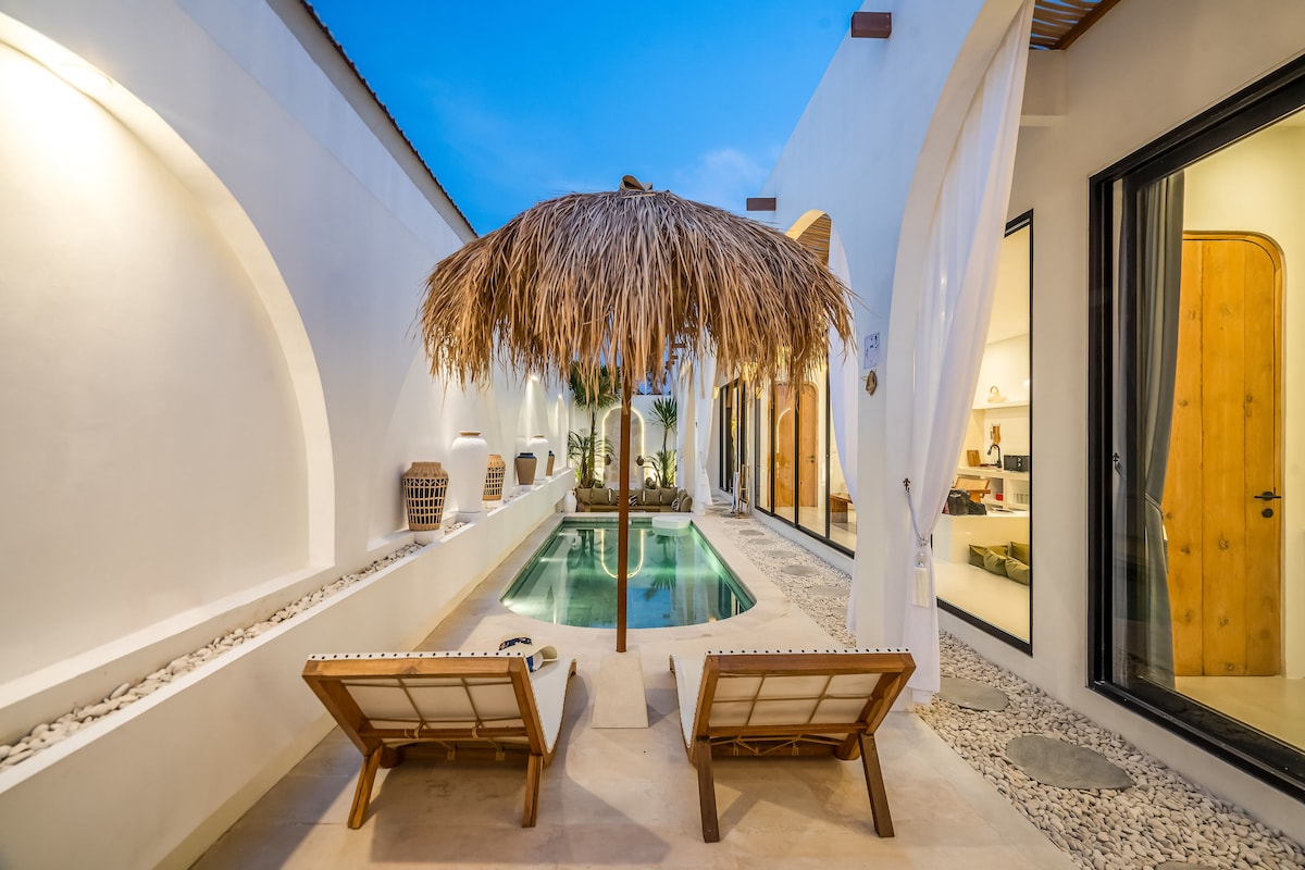 A serene outdoor space is highlighted by a thatched umbrella providing shade over two sun loungers, positioned beside a tranquil pool. The white walls and stone pathway create a calming ambiance, while lush greenery adds a touch of nature to the inviting setting.