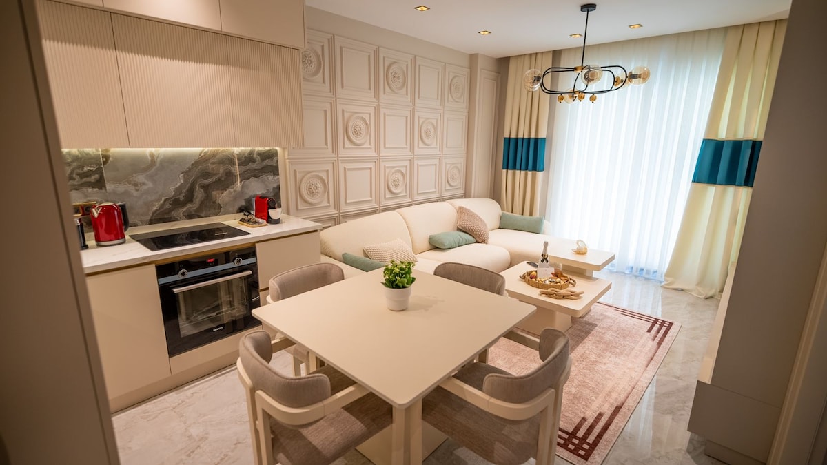 A modern living area features a light-colored sofa adorned with pastel cushions. A minimalist dining table sits nearby, complemented by plush chairs. The kitchen area includes sleek cabinets and appliances, while natural light filters through elegant drapes, enhancing the serene atmosphere.