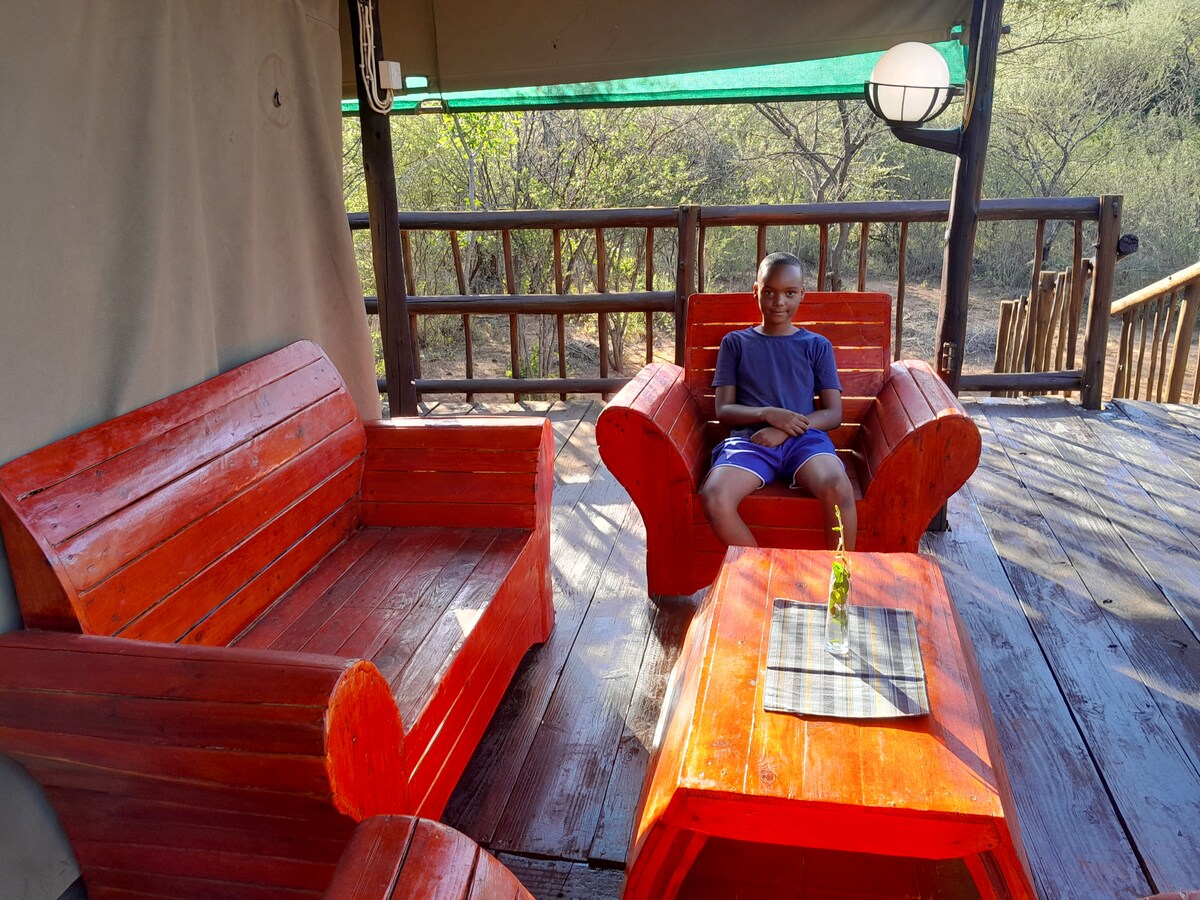 A wooden deck area features two vibrant orange chairs and a matching table, inviting relaxation. Natural greenery surrounds the space, enhancing its connection to the outdoors. A shaded cover provides comfort while sitting, with a simple centerpiece placed on the table.