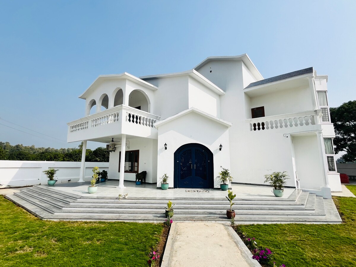 A modern farmhouse exterior is framed by lush green grass and colorful flower pots. The multi-level structure features elegant balconies and arched doorways. A spacious stone pathway leads to a large blue entrance, enhancing the inviting appeal of the property.