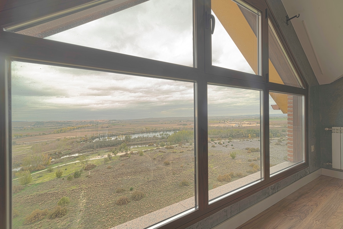 Large windows provide expansive views of the surrounding landscape, highlighting the river and rolling hills. Soft natural light fills the space, enhancing the serene atmosphere. The angular roof complements the modern design, creating a welcoming and open environment.