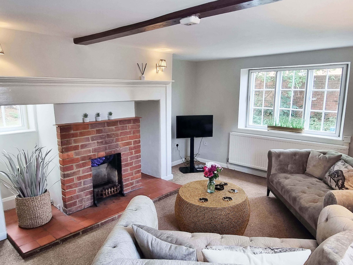 A bright living room features a brick fireplace as a focal point, flanked by a large window enhancing natural light. Comfortable seating is arranged around a woven coffee table, complemented by a floral arrangement. Additional television stands on a modest console against the wall.