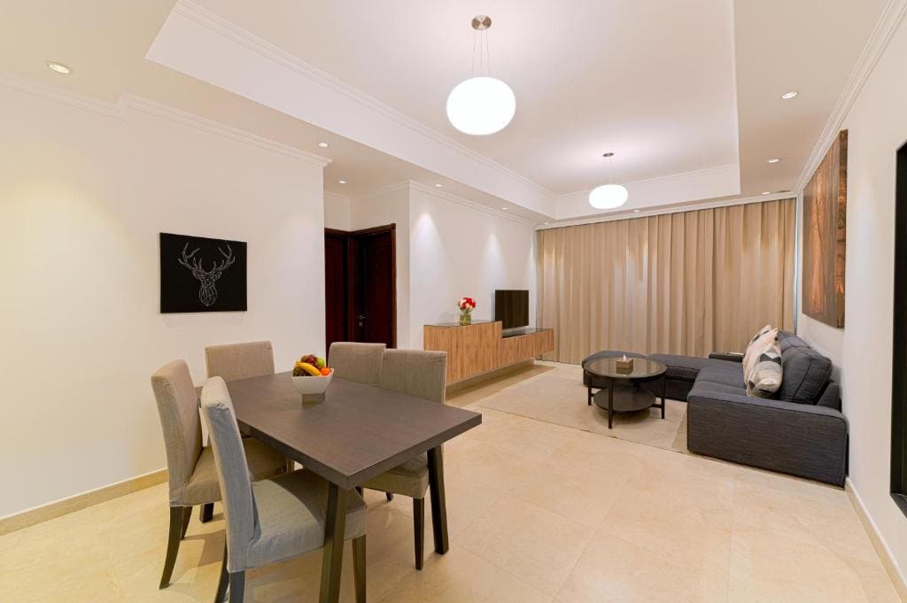 A spacious living area features a comfortable sofa and a coffee table, illuminated by overhead lighting. A dining table is positioned nearby, accompanied by upholstered chairs. Floor-to-ceiling curtains provide a soft backdrop, and a television is mounted on a minimalist cabinet.