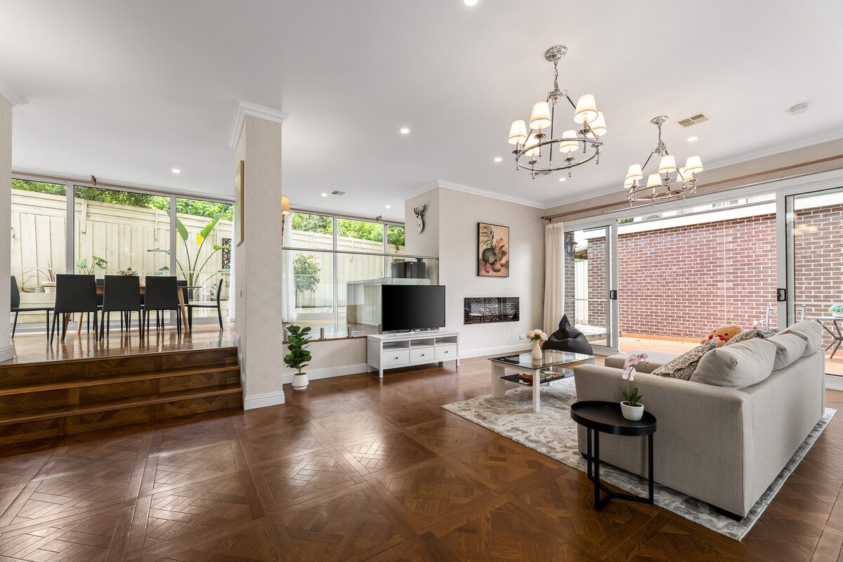 The spacious living area features a modern sofa and a flat-screen TV, complemented by an electric fireplace. Full-height windows provide views of the outdoor terrace, where a dining table awaits. A warm glow from stylish light fixtures enhances the welcoming ambiance.