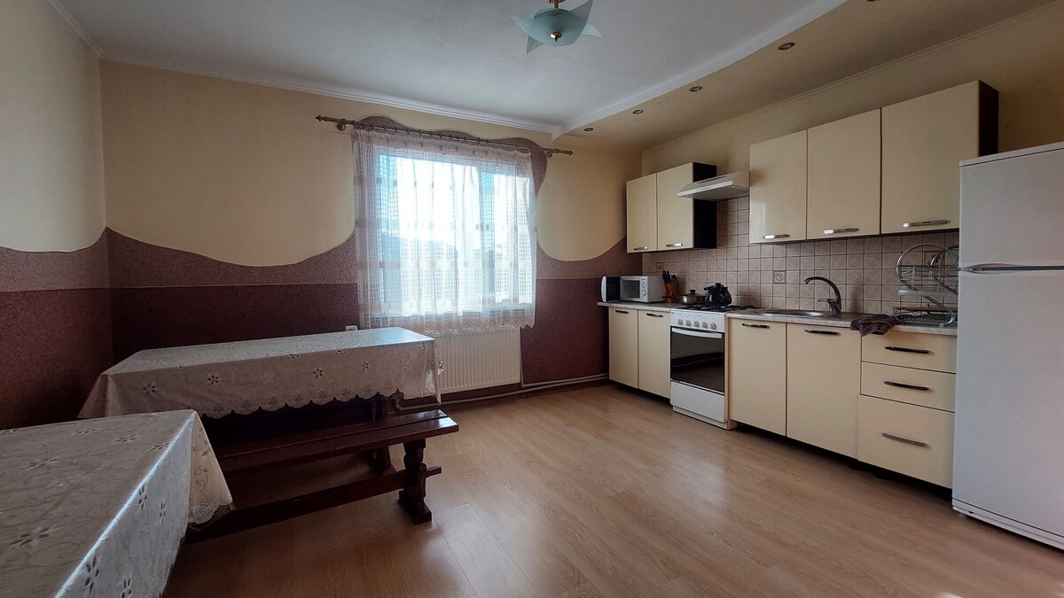 A spacious kitchen is equipped with light-colored cabinets and a large window, allowing natural light to fill the area. A dining table with benches is present, accompanied by a stove, oven, microwave, and refrigerator, providing ample space for meal preparation and dining.