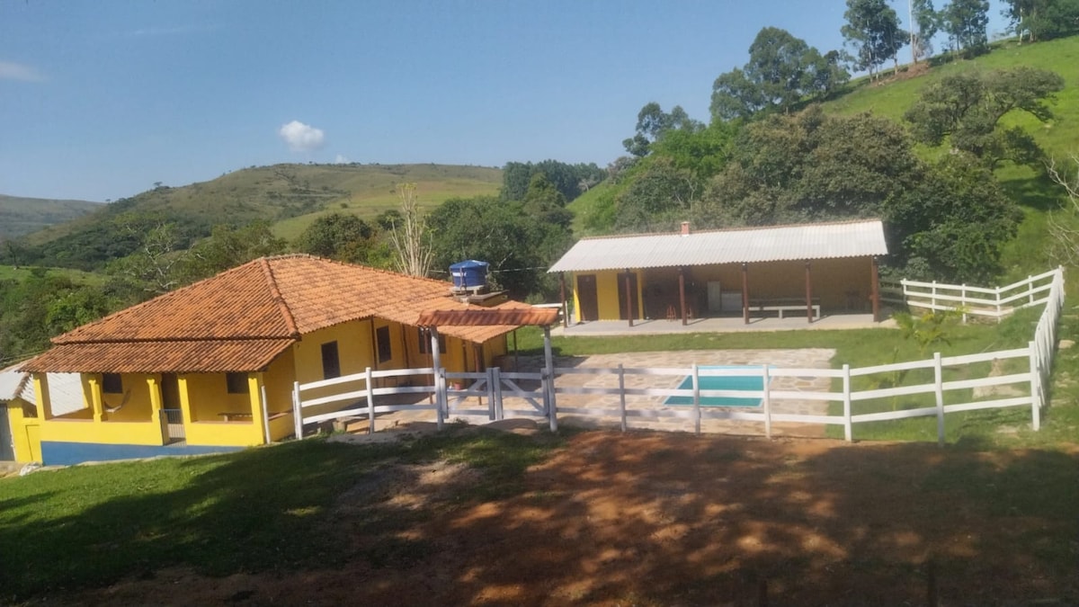 A charming country house features a bright yellow exterior with a terracotta roof. Nearby, a covered area offers seating, while a clear blue pool is situated in the outdoor space. Lush green hills and trees surround the property, enhancing its natural setting.