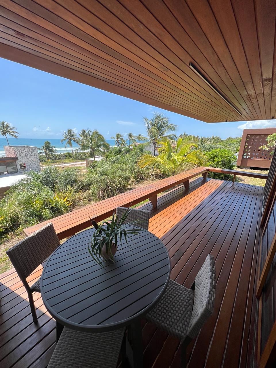 A wooden deck area is shown with a round table and four chairs, surrounded by vibrant greenery. In the background, a view of the ocean and palm trees is visible under a clear blue sky, creating a serene outdoor atmosphere.