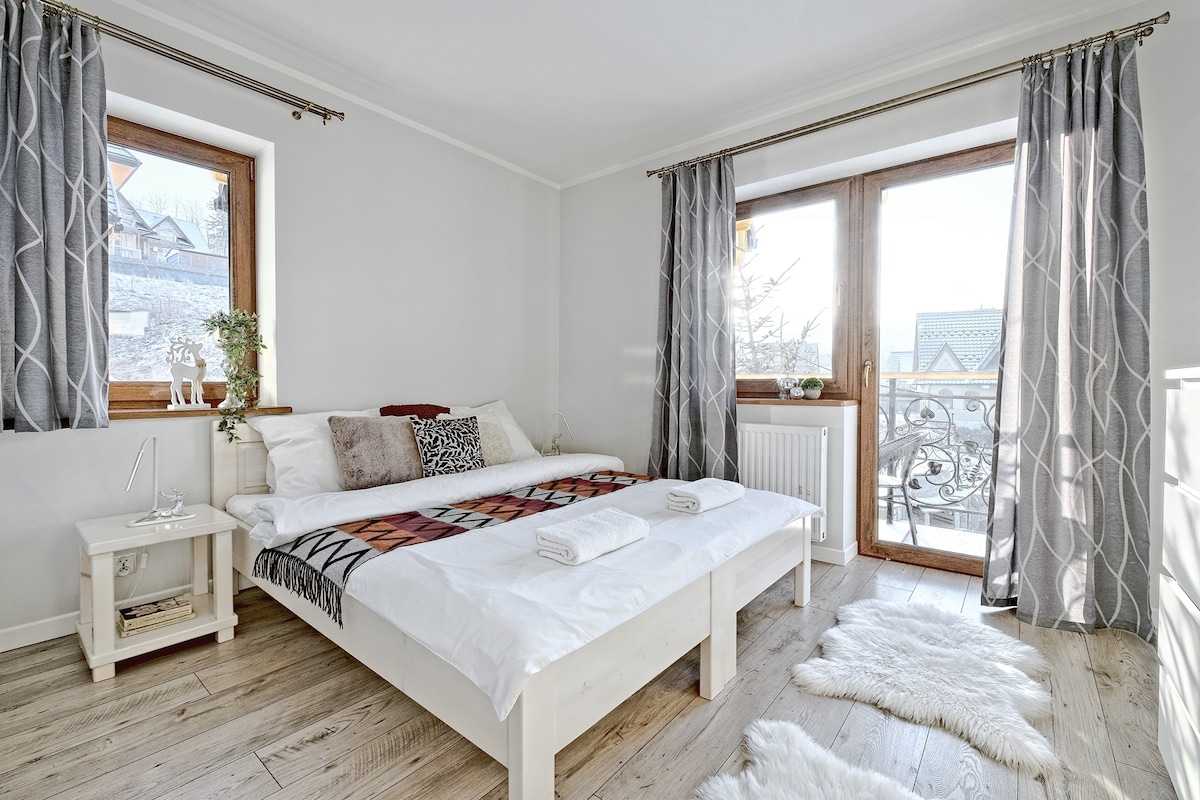 A bright sleeping area features a spacious bed accented with a patterned throw. Natural light fills the room through large windows with gray curtains. A soft rug is placed on the wooden floor, and decorative plants add a touch of nature.
