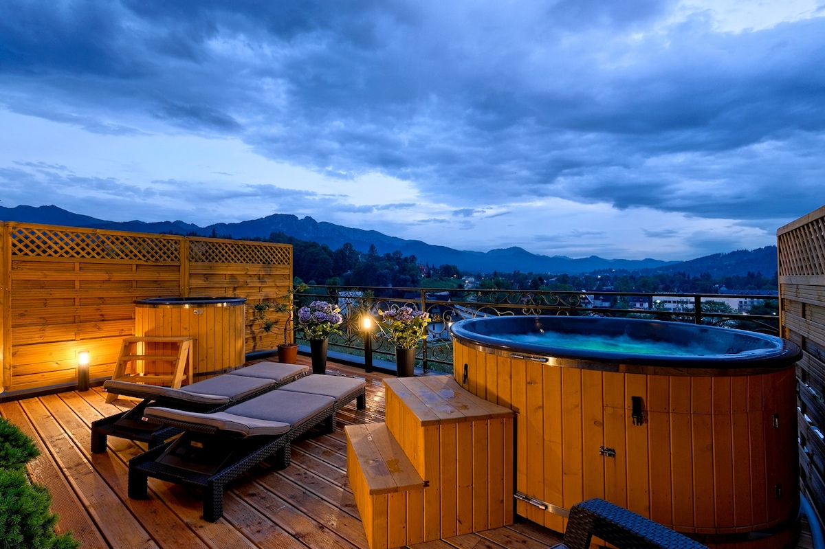 An outdoor terrace features a wooden hot tub surrounded by seating areas and potted plants. Soft lighting enhances the ambiance against a backdrop of mountains under a cloudy sky, creating a serene spot for relaxation.
