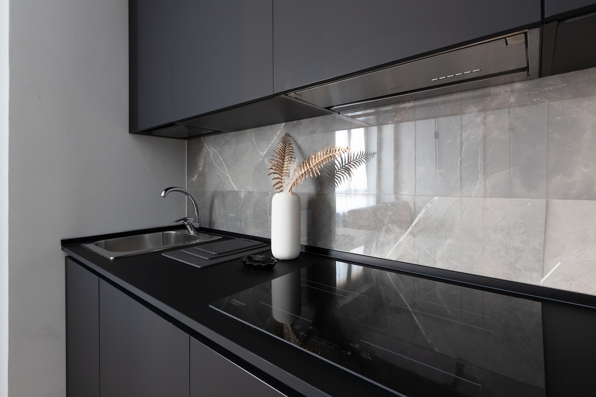 A modern kitchen area features sleek dark cabinetry and a minimalist design. A stainless steel sink is positioned next to an induction cooktop, with a decorative white vase holding a dried fern on the counter, complemented by a polished marble backsplash.