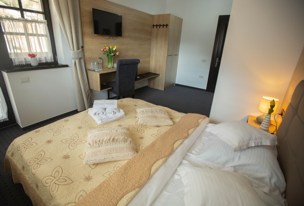 A well-appointed bedroom features a spacious bed covered with a light-colored throw. Neatly folded towels are placed on the bed. A sleek desk with a chair and a television mounted on the wall enhance the functionality of the space, while natural light filters through the window.