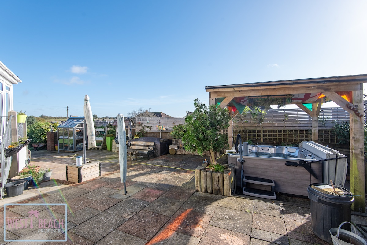 An outdoor area is shown, featuring a hot tub under a shaded gazebo. Paved stone flooring is visible, along with planters and a garden area. A few deck chairs and an umbrella can be seen, creating an inviting space for relaxation.