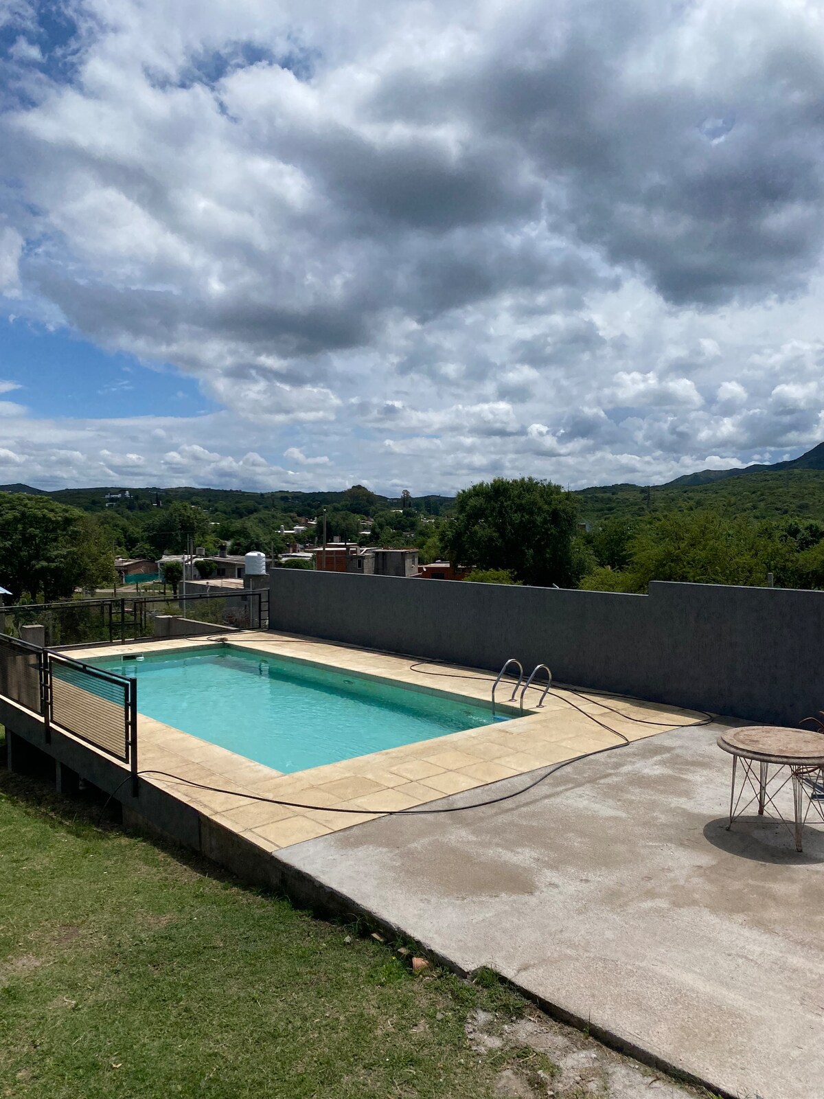A clear swimming pool is positioned within a spacious outdoor area, surrounded by a sun terrace and low fencing. The view encompasses green hills and a small town in the distance, with partly cloudy skies overhead.