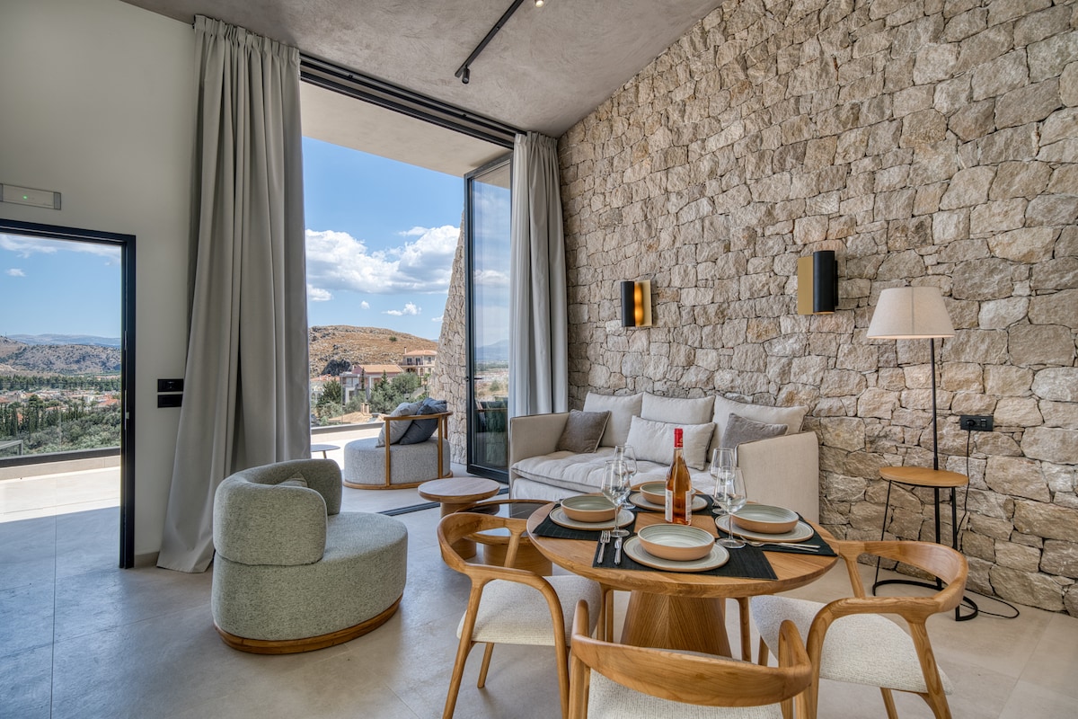 A spacious living area features a light-colored stone wall, complemented by large glass doors that open to outdoor views. A round dining table is set for four, while a comfortable sofa and a modern accent chair invite relaxation. Natural light brightens the ambiance.