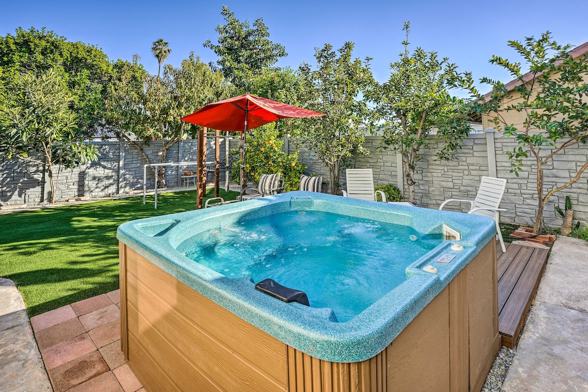 A private hot tub is surrounded by a landscaped backyard, featuring green grass and fruit trees. Sun loungers are positioned nearby under a shaded canopy. The bright blue sky creates a serene atmosphere, enhancing the inviting outdoor setting.