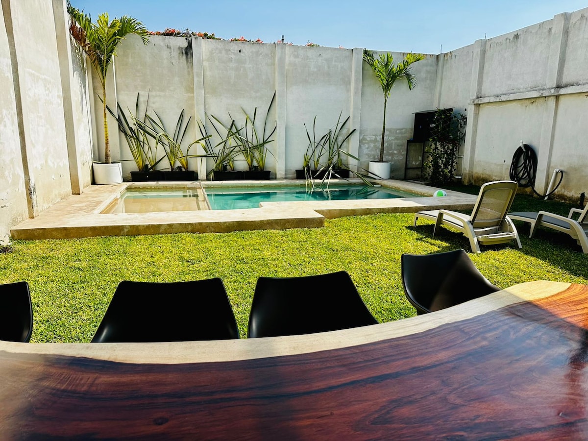 A manicured garden surrounds a rectangular, climatized pool, inviting relaxation. Sun loungers are positioned nearby, and a spacious wooden table, crafted from natural wood, is seen in the foreground, offering a perfect spot for gathering. Vertical plants add greenery against the white walls.