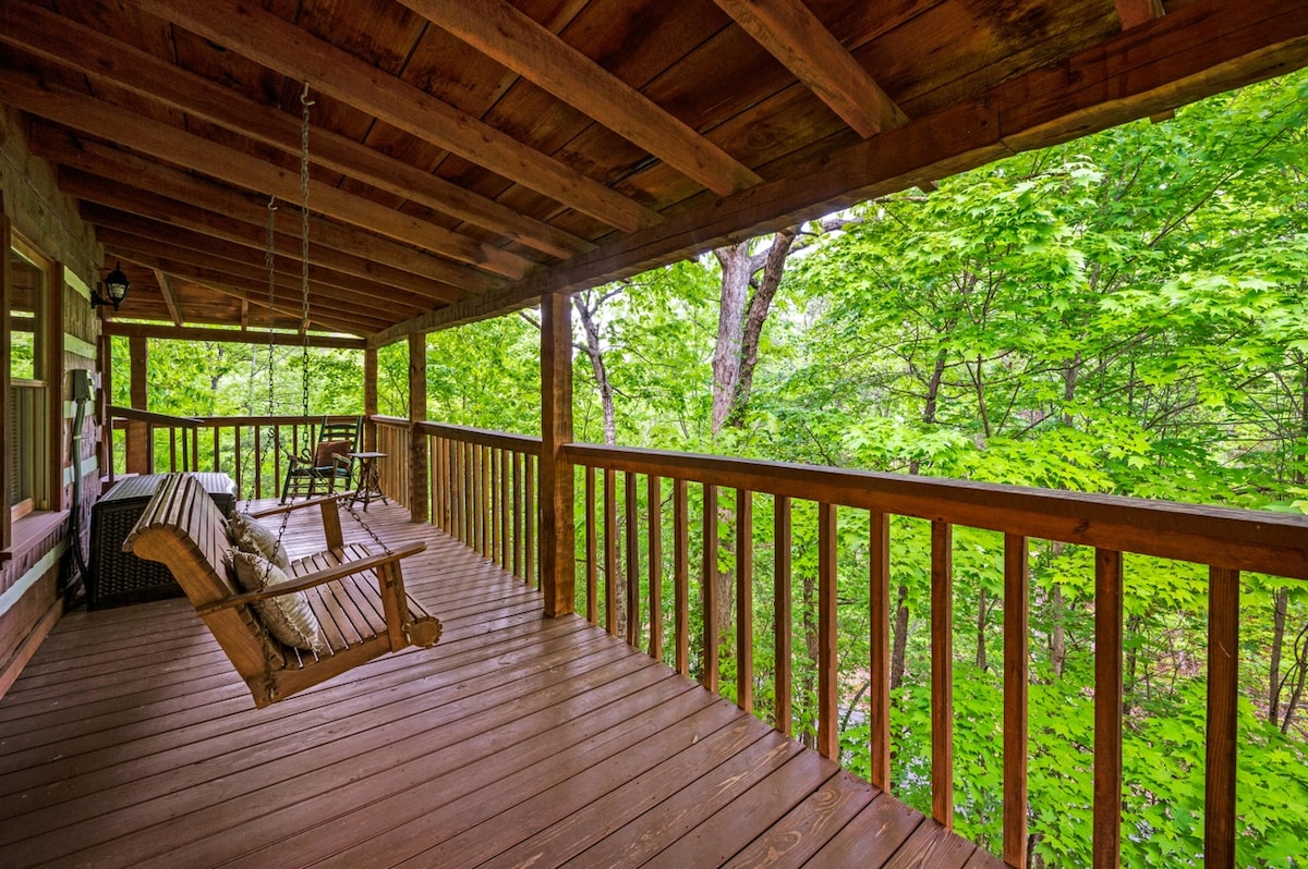 A spacious wooden porch features a swing and a couple of chairs, surrounded by lush greenery. Sunlight filters through the leaves, creating a tranquil setting for relaxation. The natural wood tones of the porch enhance the connection to the serene forested environment.