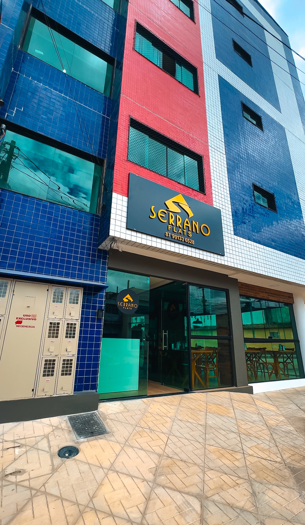 The exterior of the building features a vibrant façade composed of blue and red tiles. The entrance is marked by a large sign that reads 'SERRANO.' Large windows allow light to flow into the common areas, and nearby, a row of lockers can be seen.