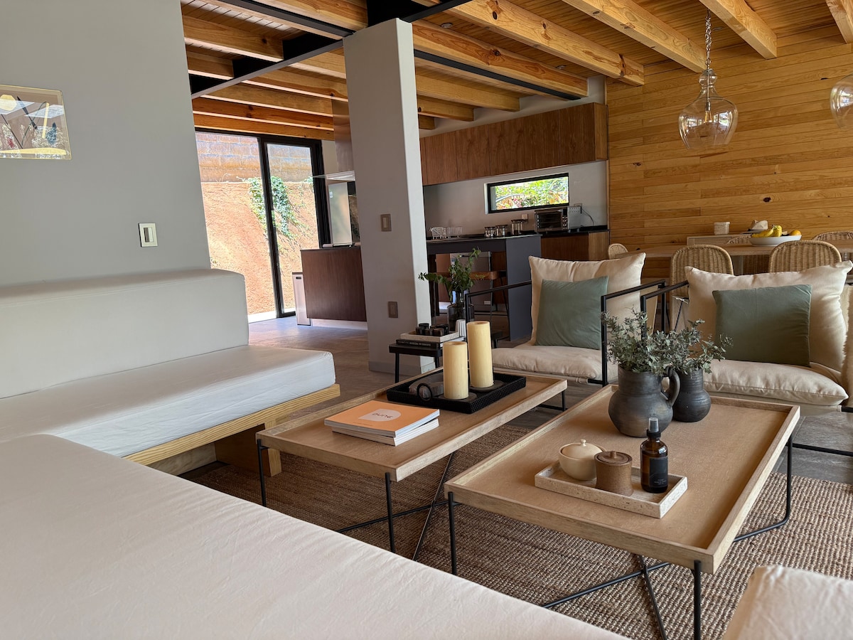 A modern living space is showcased with natural wood beams overhead and a light color palette. Comfortable seating is arranged around a coffee table that features decorative elements, candles, and a small vase with greenery. An open kitchen area is partially visible in the background.