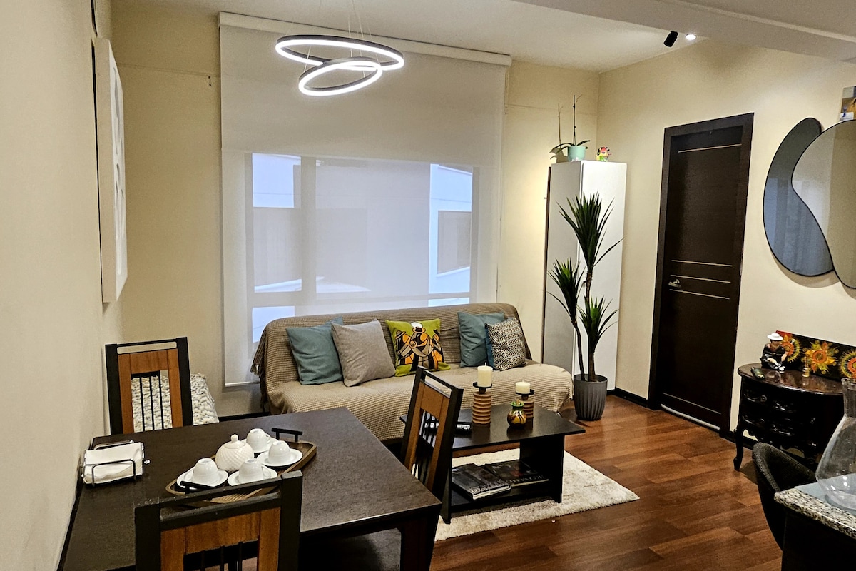A contemporary living space features a light-colored sofa adorned with decorative pillows, accompanied by a dining table and four chairs. A floor plant adds a touch of greenery, while natural light filters through the sheer window coverings. Modern light fixtures are positioned above the dining area.