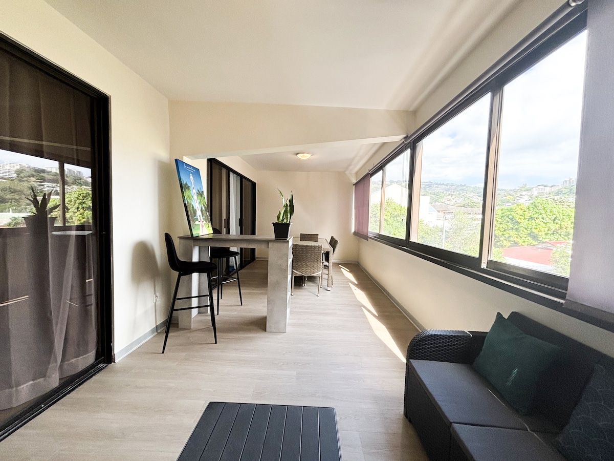 A bright and airy terrace features large windows allowing natural light to fill the space. A sleek bar table with stools stands at one end, while comfortable seating is arranged on the opposite side. Greenery is visible outside, enhancing the peaceful ambiance.