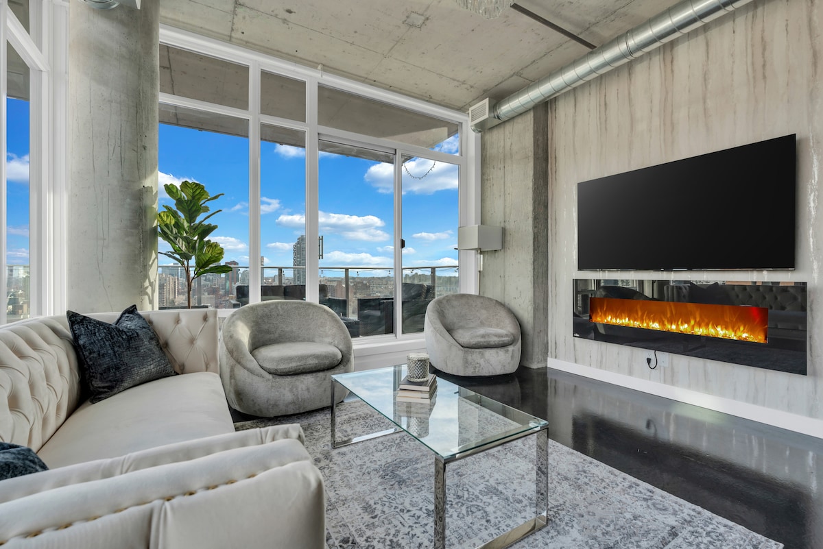 A spacious living area features floor-to-ceiling windows that showcase panoramic city views. Plush seating is arranged around a glass coffee table, while an electric fireplace is mounted on the wall beneath a large television. Natural light floods the room, creating a bright atmosphere.