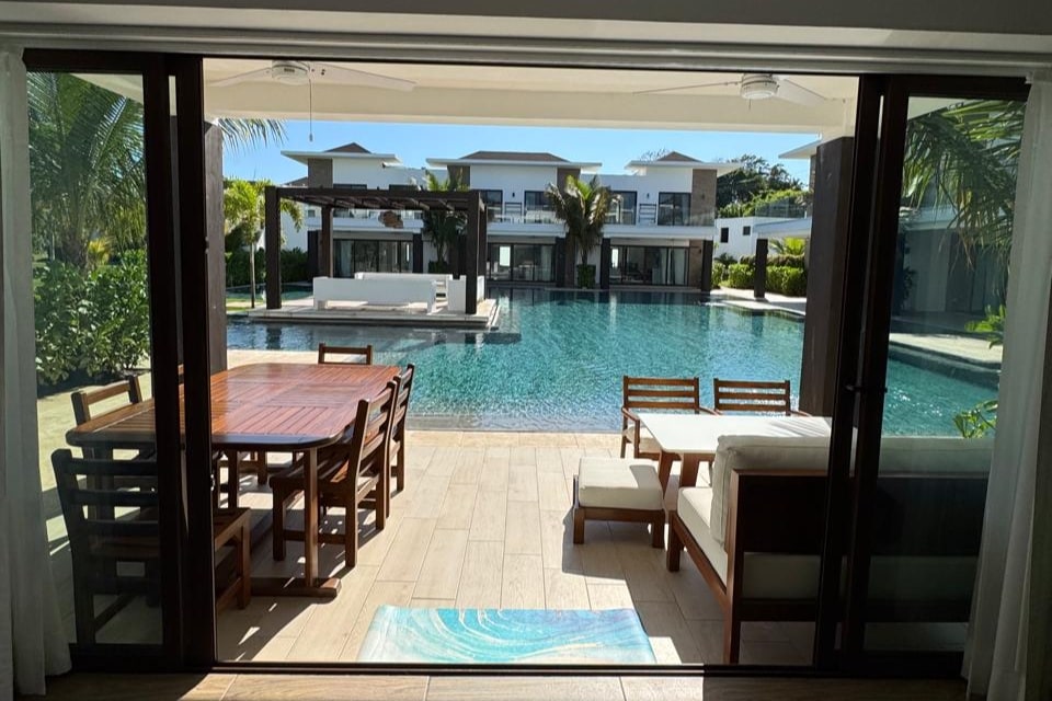 An outdoor dining area is visible, featuring a wooden table surrounded by several chairs. Beyond, a large, serene swimming pool reflects the sunlight, bordered by tropical greenery. Smooth glass doors create a seamless connection between the indoor and outdoor spaces.