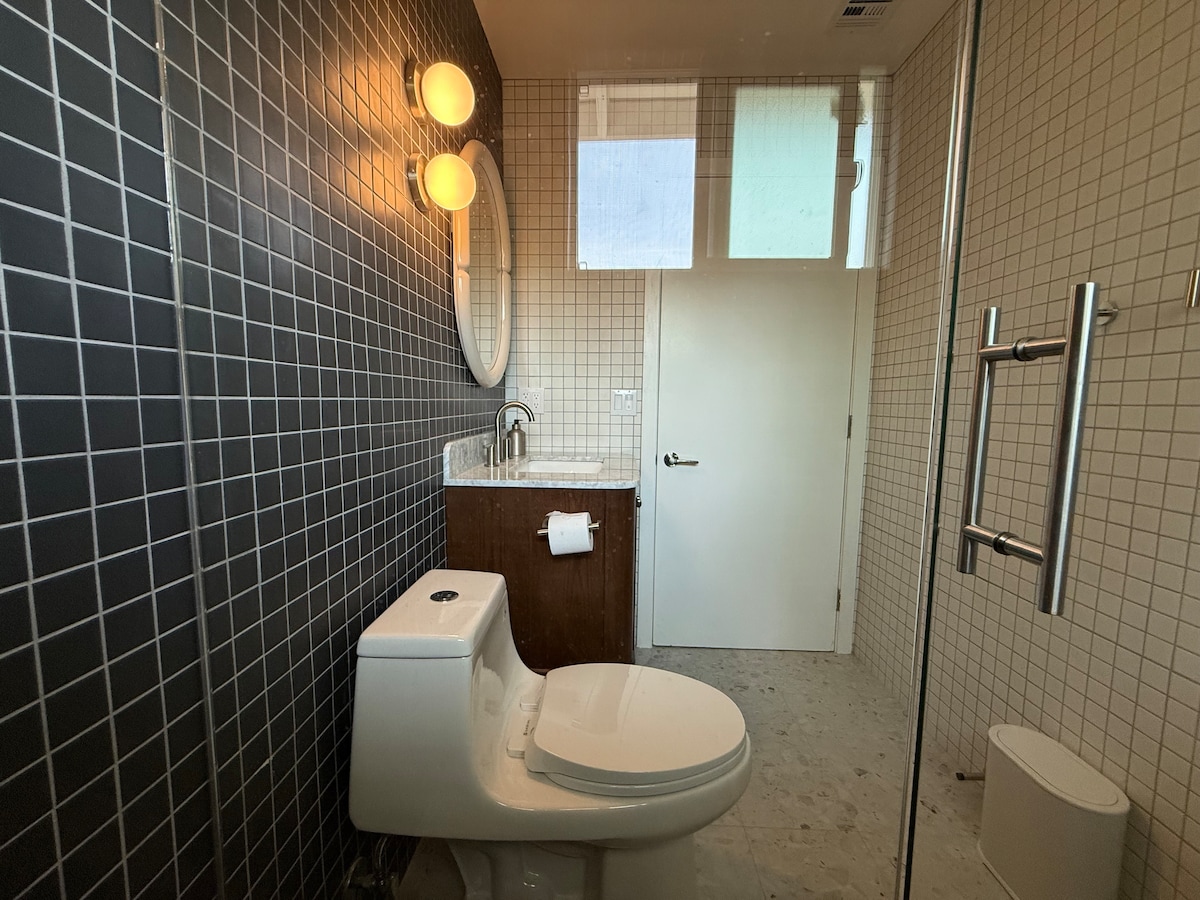 A modern bathroom is presented with tiled walls in a grid pattern. A spacious shower stall is visible, accompanied by a sleek toilet and a wooden vanity with a stone countertop. Two round light fixtures provide illumination, and frosted glass windows allow natural light while ensuring privacy.