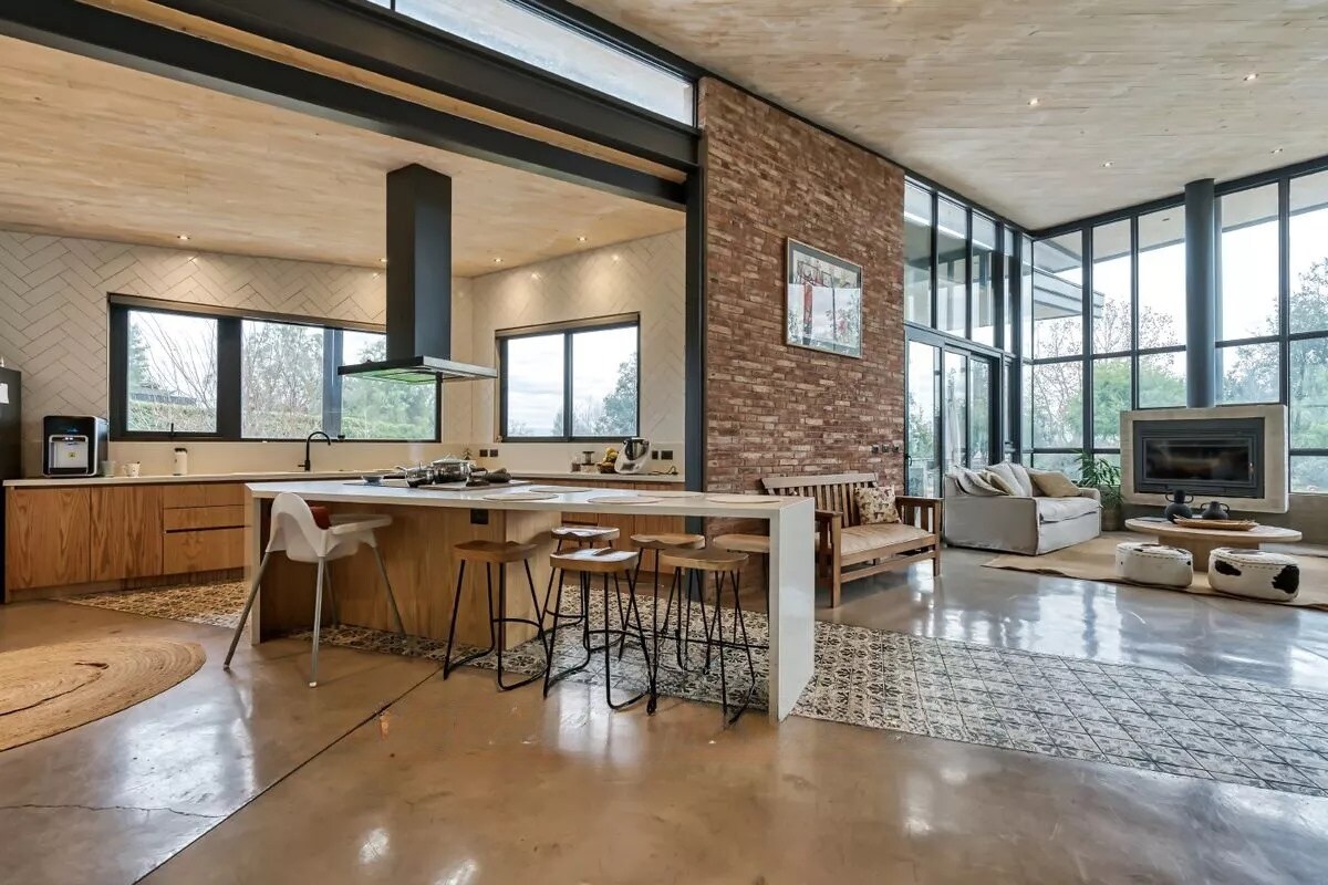 An open-concept living area features a modern kitchen with a large island, accompanied by wooden stools. A comfortable seating area is defined by large windows, allowing natural light to fill the space. A cozy fireplace and natural textures enhance the inviting ambiance.