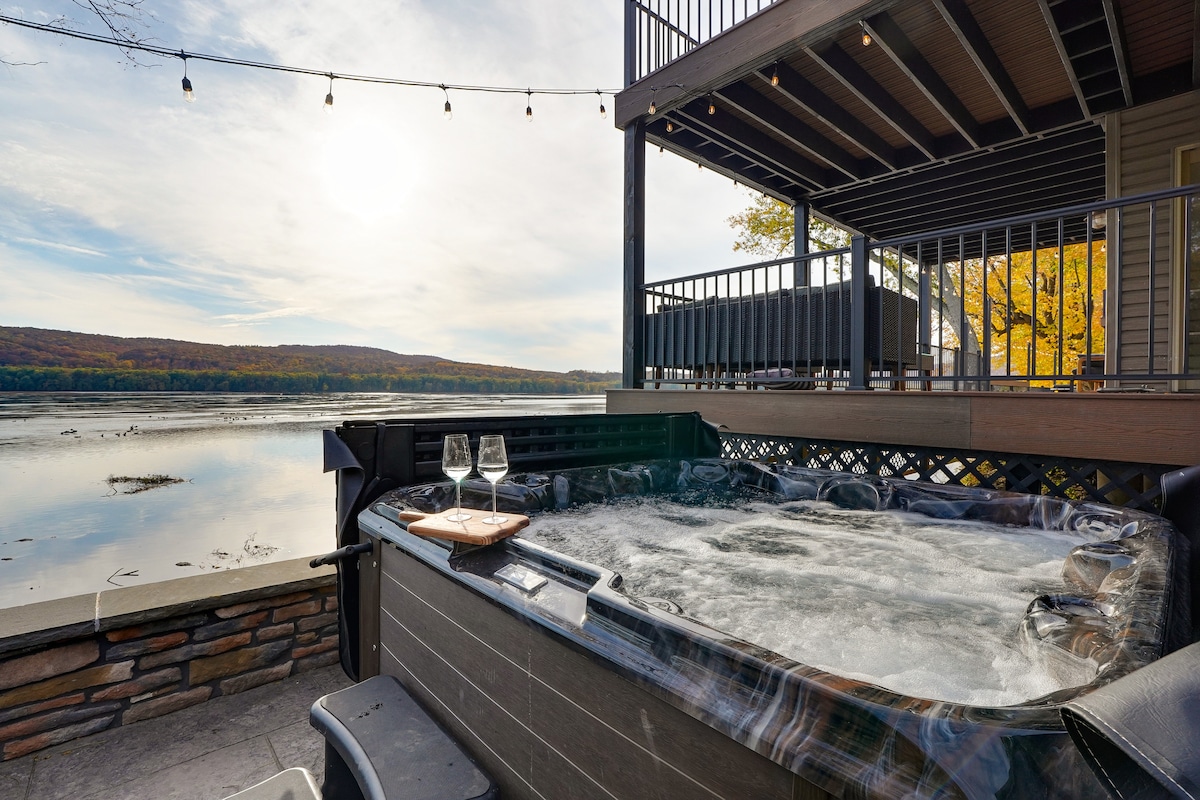 The hot tub is positioned on the patio, overlooking the Susquehanna River. Two glasses sit on a wooden tray beside the tub, while string lights create a soft glow above. Surrounding trees show fall colors, and the sun sets in the background, reflecting on the water.