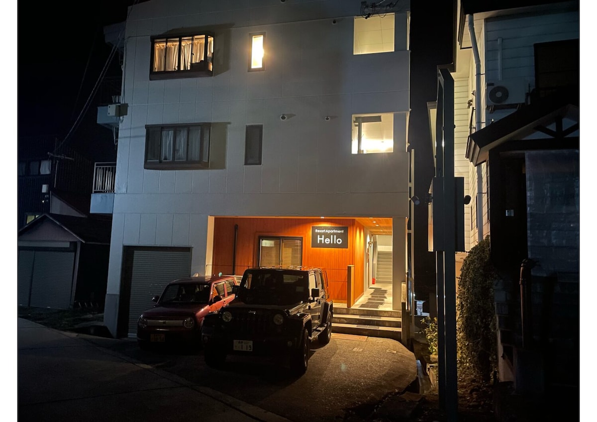 An illuminated entryway stands at the forefront of a modern building at night. Large windows allow light to spill out, while a welcoming sign reads 'Hello.' Vehicles are parked in the driveway, showcasing the building’s accessibility.
