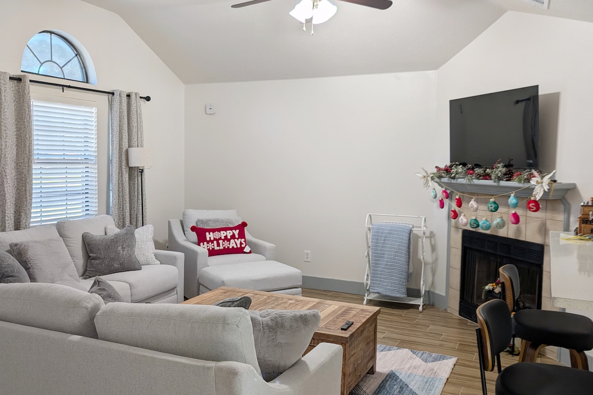 A bright living area features a light-colored sectional sofa and an accent chair. A wooden coffee table sits at the center, with a television mounted on the wall above a decorated mantel. Holiday decor adds a seasonal touch to the space.