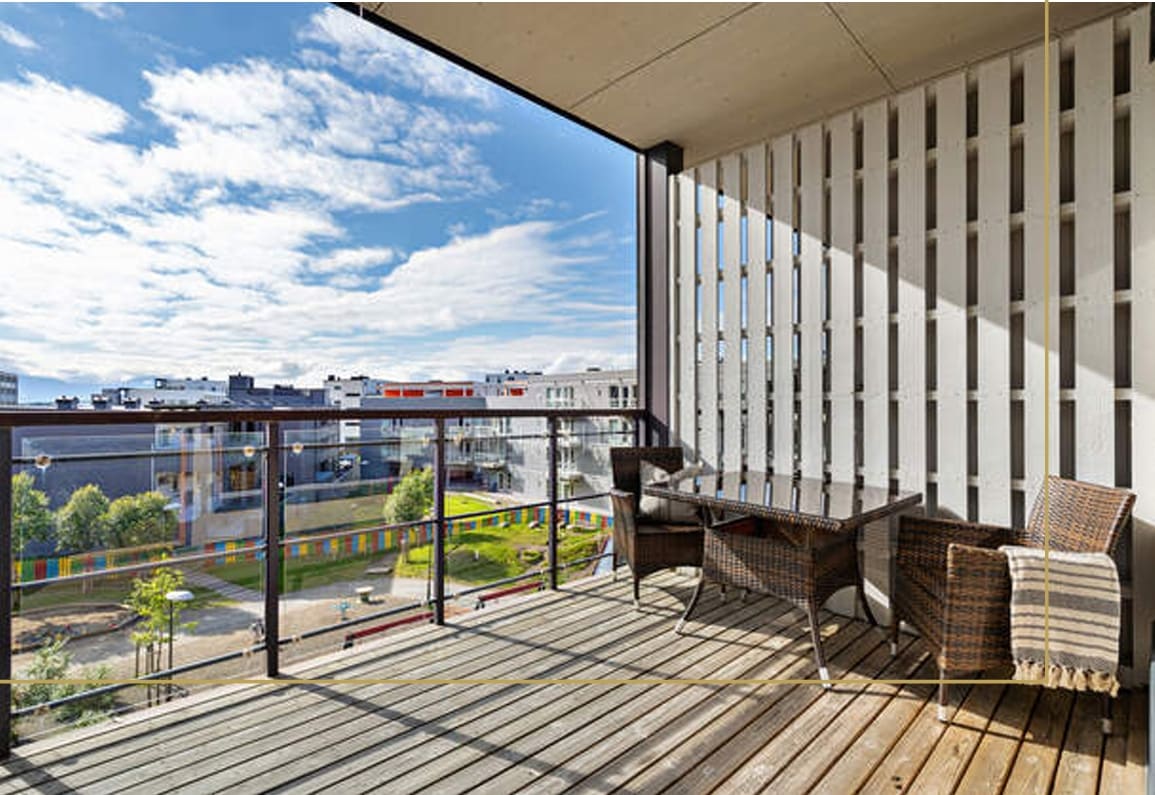 A spacious balcony features woven furniture, including a table and two chairs. It overlooks a landscape with well-maintained greenery and colorful structures. Soft sunlight filters through the slatted walls, creating contrasting shadows on the wooden deck.