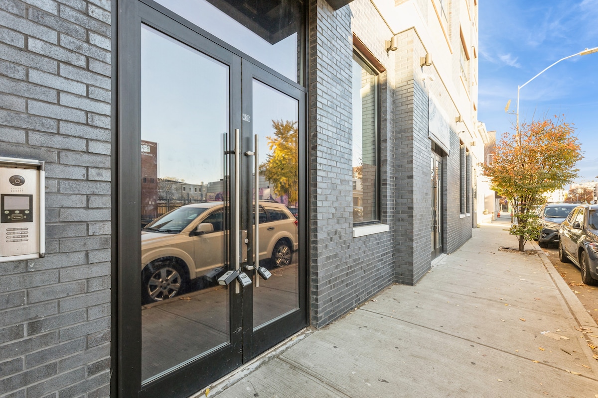 The entrance features a modern design with large glass doors framed in sleek dark materials. A nearby intercom and mailbox area can be seen, along with a clean sidewalk and street lined with vehicles and a tree displaying autumn foliage.