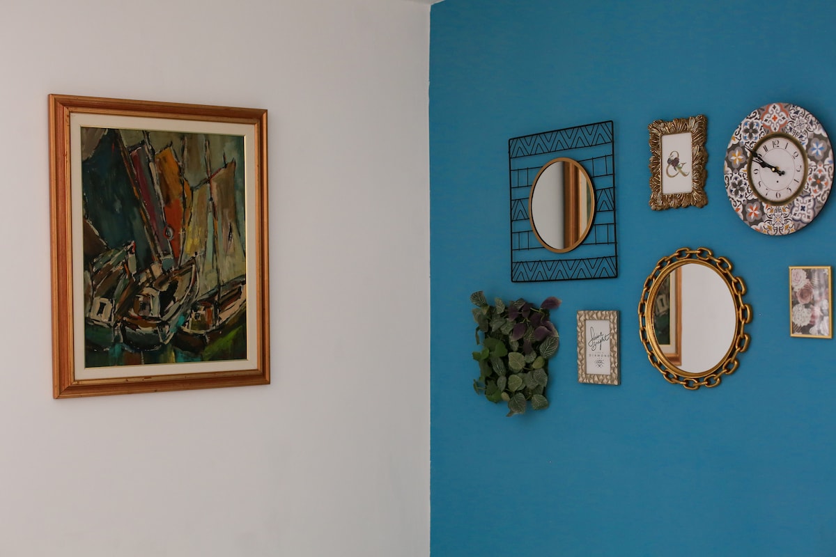 A collection of decorative wall items enhances the living space. An abstract painting of boats is displayed in a wooden frame, complemented by several mirrors in varied shapes and sizes, alongside a printed artwork and a small plant draped in a corner.