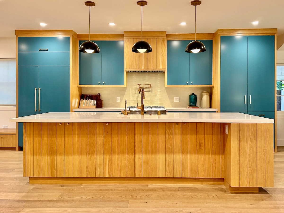 The kitchen features a spacious island topped with light quartz, surrounded by custom cabinetry in deep teal and natural wood. Modern pendant lights hang from the ceiling, providing focused illumination over the cooking area. Appliances blend seamlessly into the design.