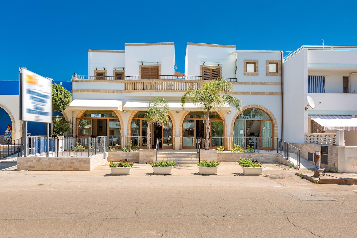The exterior of the property is prominently displayed, featuring a welcoming entrance with a series of arches. Planters with greenery and bright flowers line the front, while a clear blue sky serves as a backdrop. The building's structure showcases a mix of white and light-colored facades.