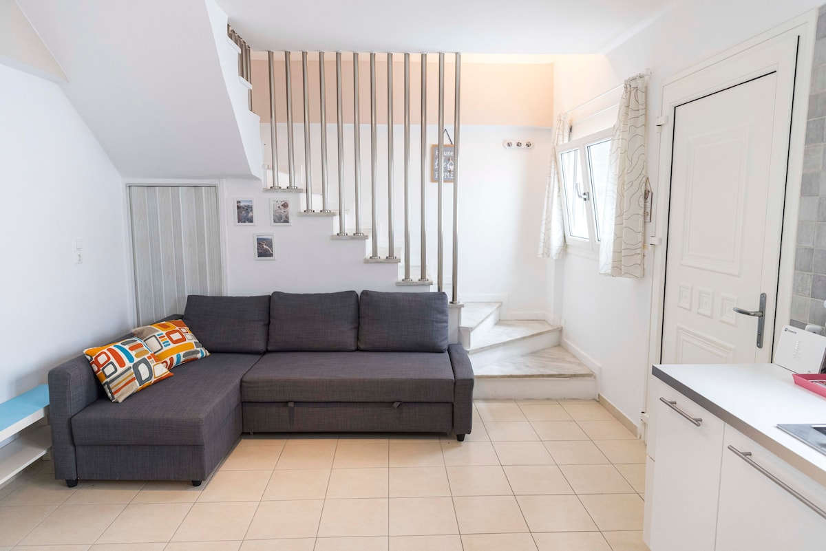 A modern living area features a gray sectional sofa with colorful patterned cushions. Natural light fills the space, highlighting tiled flooring. A staircase leads to an upper level, while light-colored walls create a bright atmosphere. Decorative framed photos are displayed on one wall.