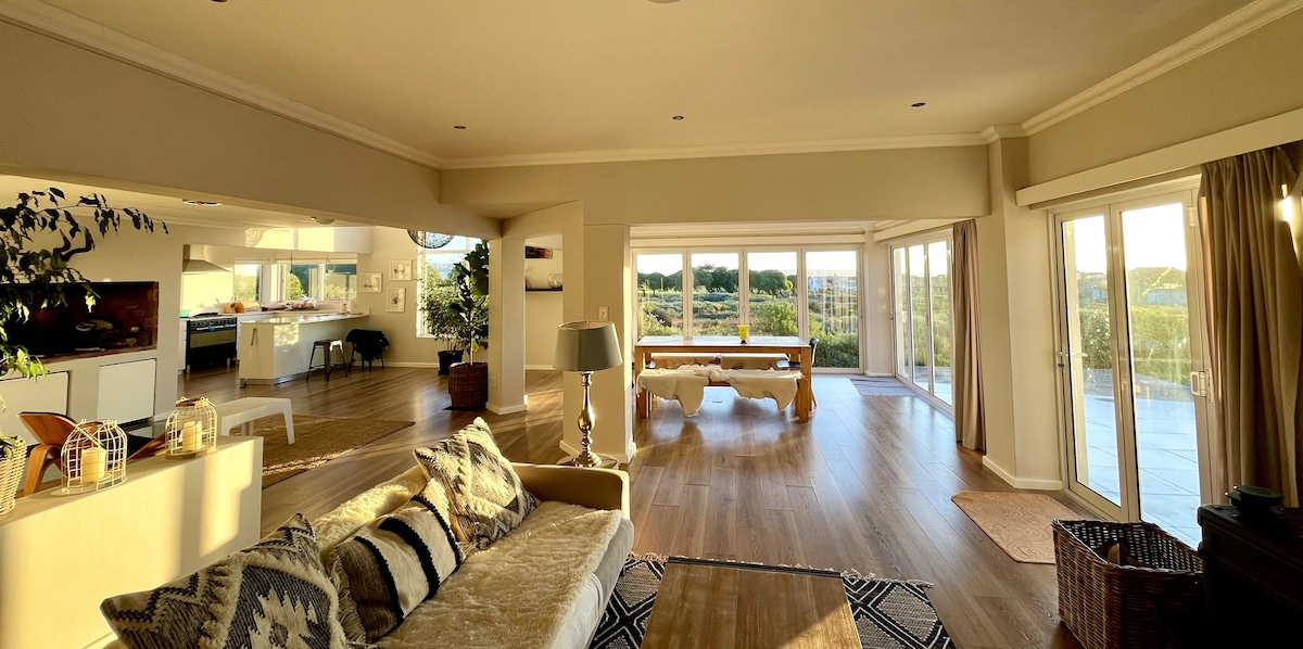 A spacious living area showcases large windows allowing for natural light, with views of greenery beyond. A comfortable sofa sits on a patterned rug, while a pool table is positioned toward the back. Soft lighting fixtures and indoor plants add to the inviting atmosphere.
