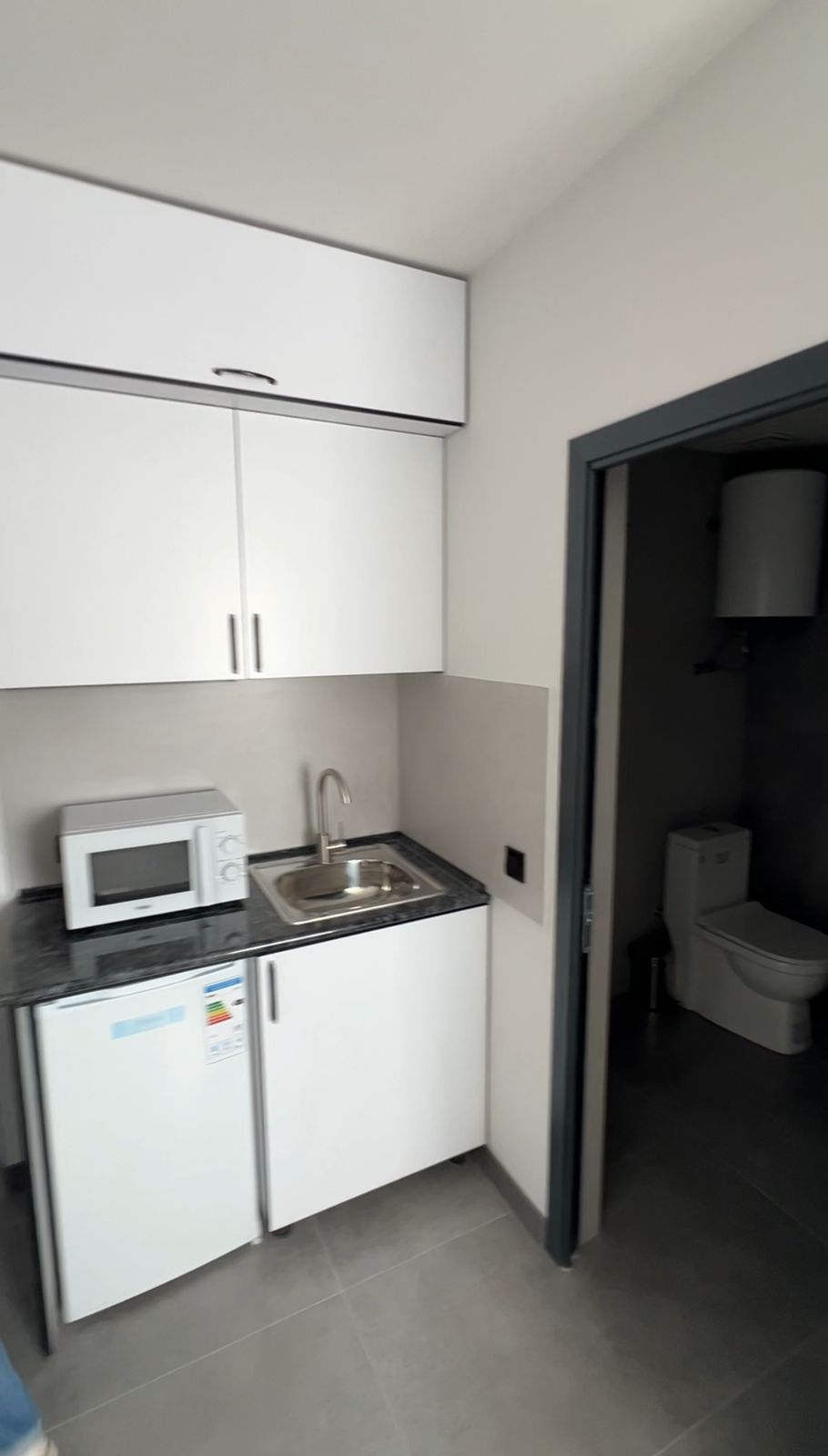 A compact kitchenette is presented, featuring a refrigerator and a microwave. A stainless steel sink is positioned next to the countertop, while cabinetry above offers storage. A doorway leads to a separate bathroom area, partially visible in the image.